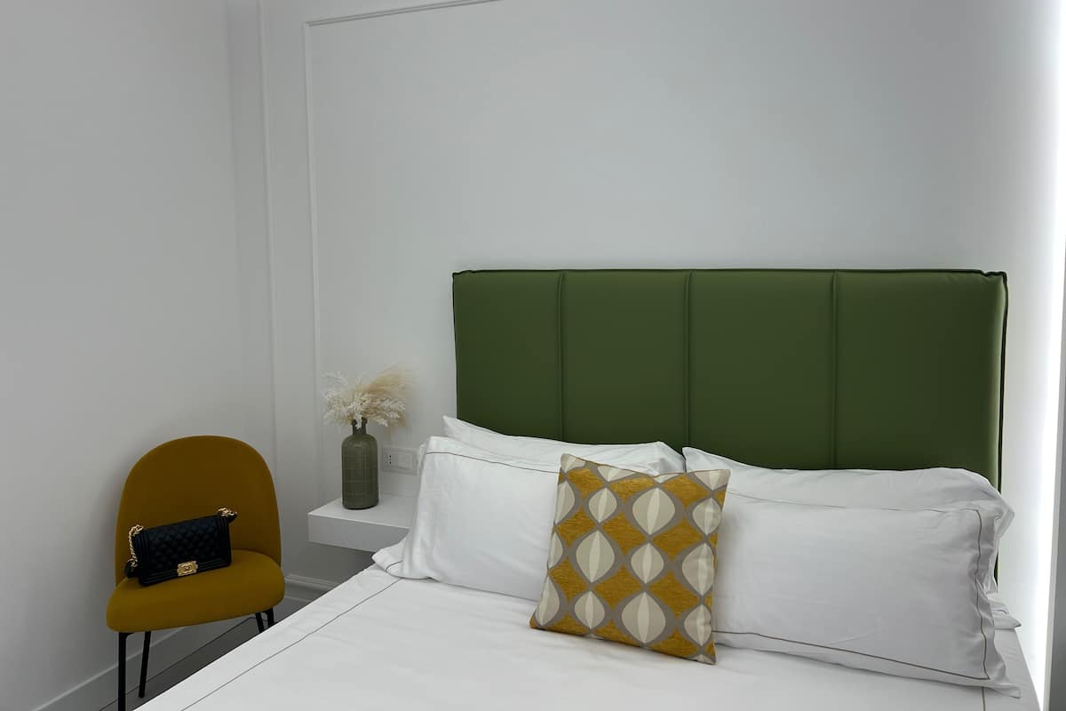 A bedroom with a green upholstered headboard, white bedding, a mustard-colored pillow, a black handbag on a mustard-colored chair, and a vase with dried flowers on a small white shelf.