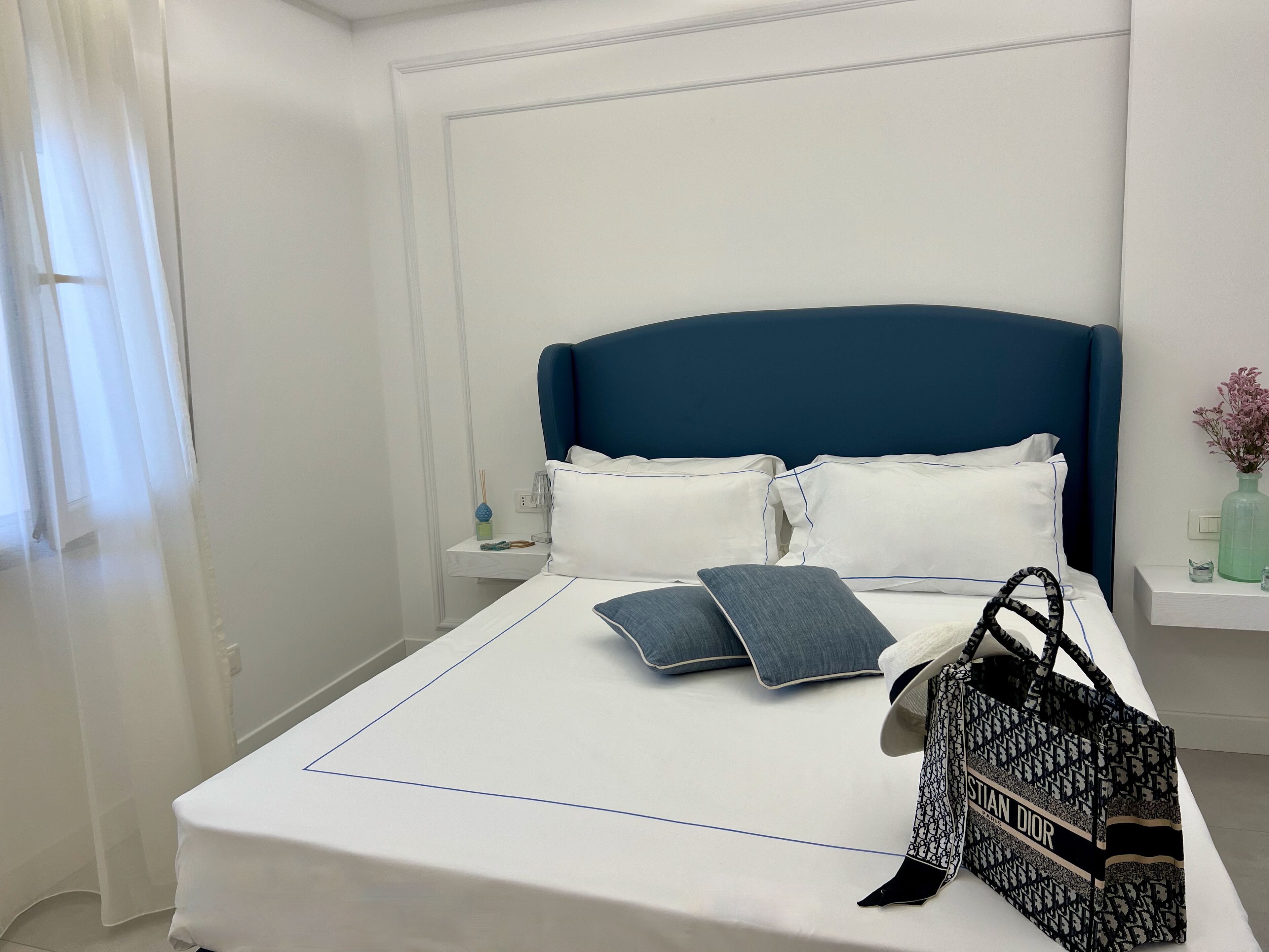 Una cama bien hecha con un cabecero azul, ropa de cama blanca y dos almohadas. Un bolso está colocado sobre la cama.