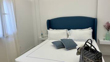 Una cama bien hecha con un cabecero azul, ropa de cama blanca y dos almohadas. Un bolso está colocado sobre la cama.