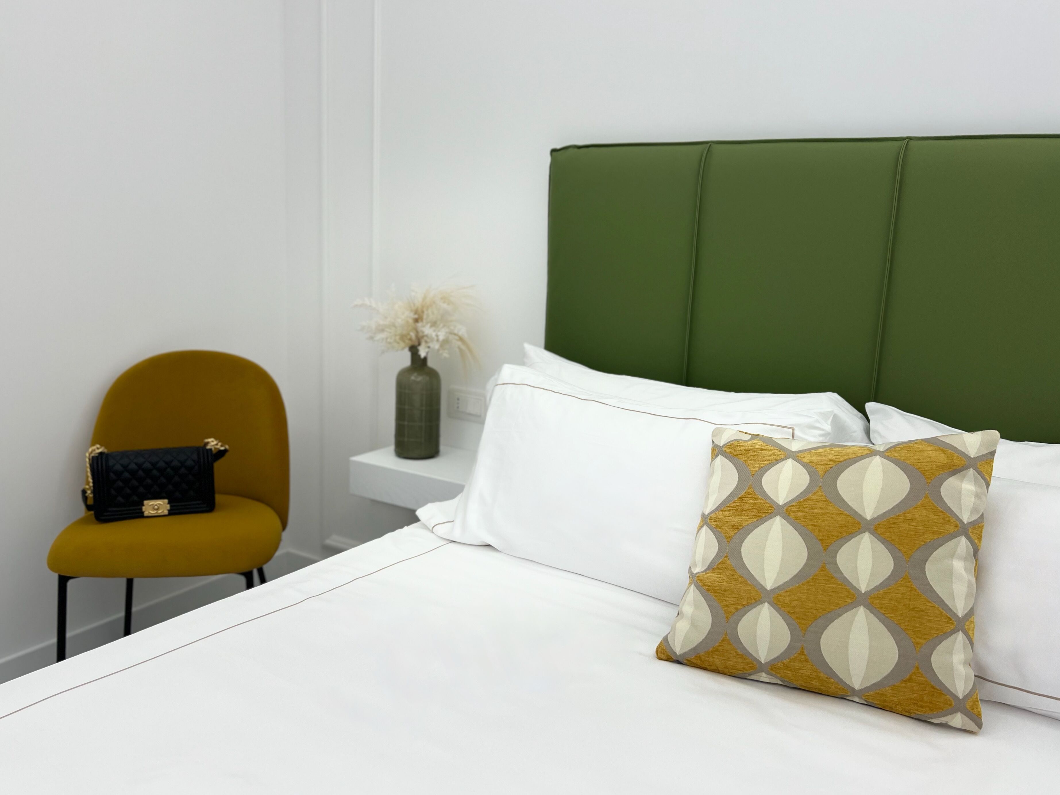 Habitación de hotel ordenada con una tapicería verde en la cabecera, una colcha blanca, un cojín estampado color mostaza, un cojín blanco y un bolso negro pequeño en una silla color mostaza.
