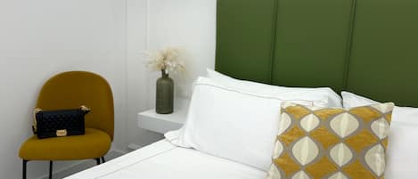 Habitación de hotel ordenada con una tapicería verde en la cabecera, una colcha blanca, un cojín estampado color mostaza, un cojín blanco y un bolso negro pequeño en una silla color mostaza.