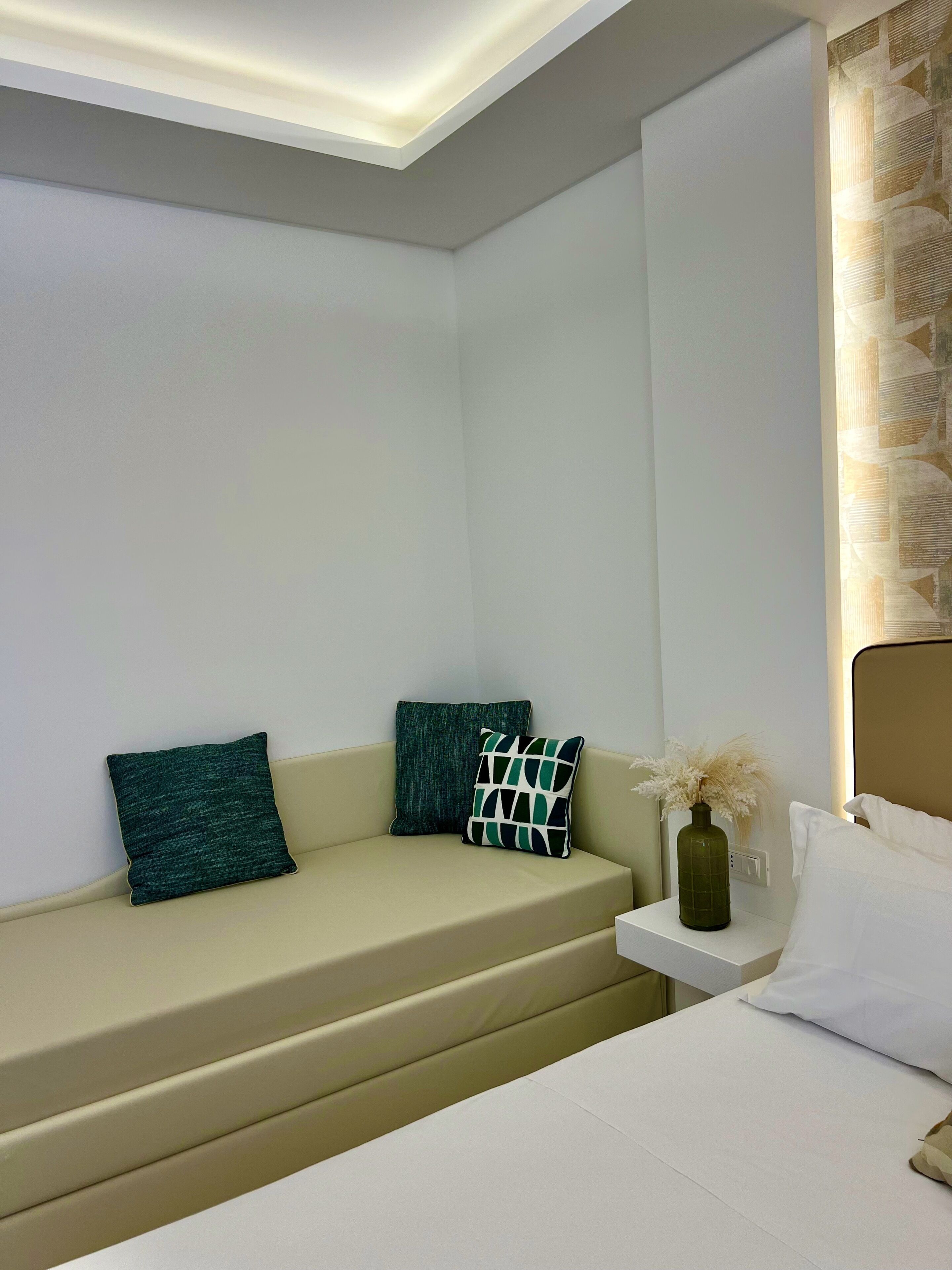 A modern hotel room with a beige sofa, two pillows, a small bedside table with a vase, and a bed with white bedding.