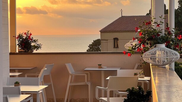 A terrace with white chairs and tables, overlooking a sunset over the sea, with a building in the background. - Kairos Tropea (Tropea)