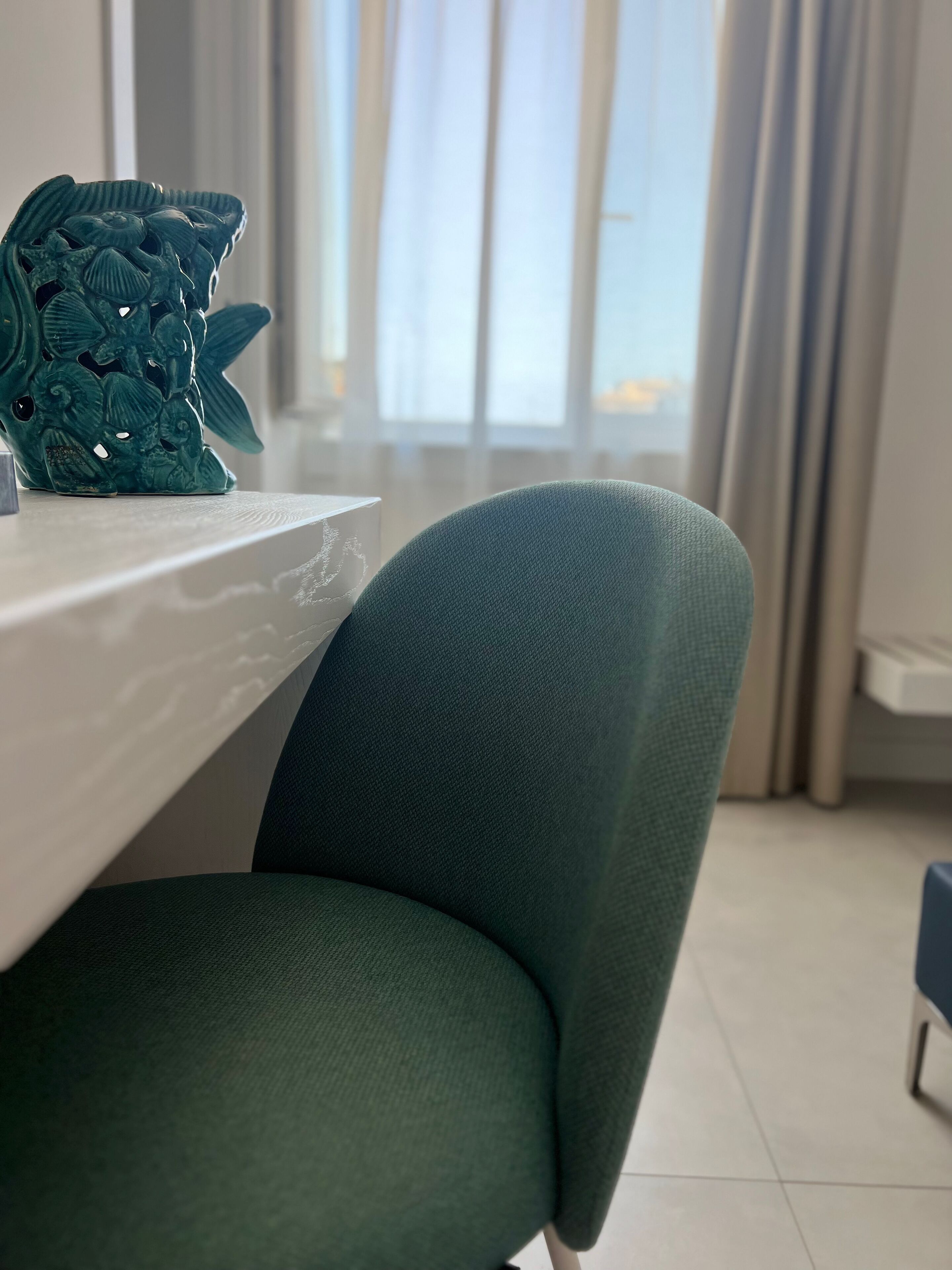 Een smaragdgroene stoel met een gestructureerde stof, een witte tafel met een decoratief object, en een raam met gordijnen in een kamer met een betegelde vloer.