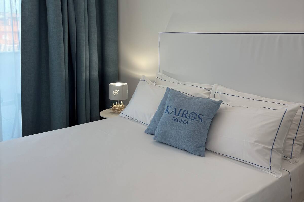 A neatly made bed with white linens and a single blue pillow with text, a bedside table with a lamp, and a window with blue curtains.