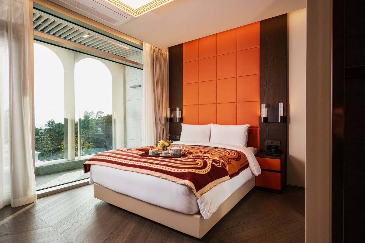 A modern hotel room with a large bed, a nightstand, a wall-mounted lamp, and a view of greenery through large windows.
