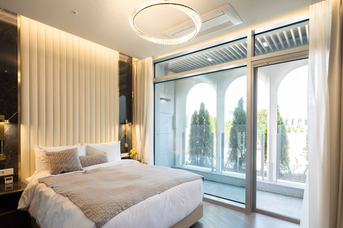 A modern bedroom with a large bed, a nightstand, a wall-mounted lamp, and a view through a glass door to the outdoors.