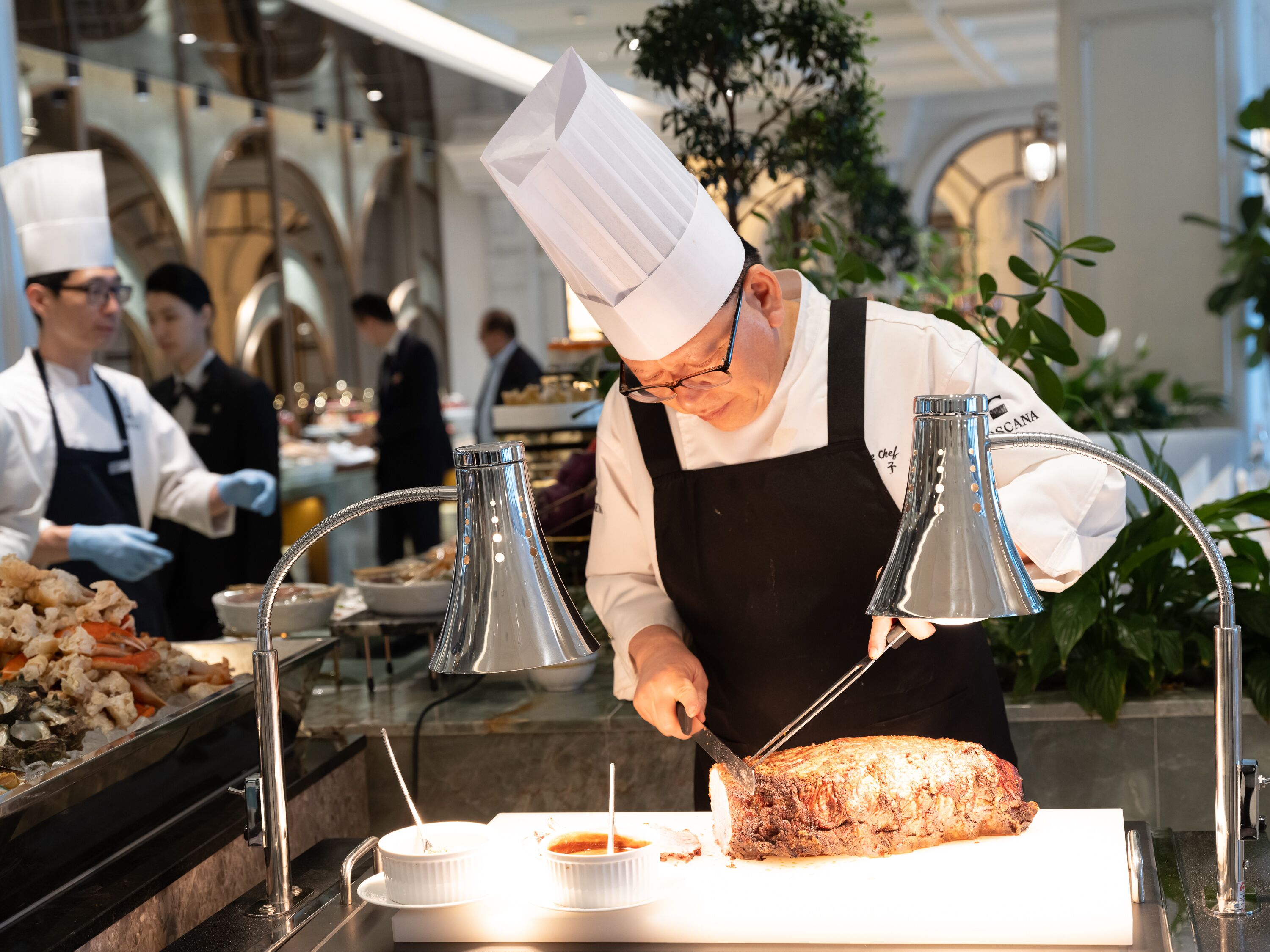 A chef slicing meat at a buffet with a modern interior and other staff members in the background.