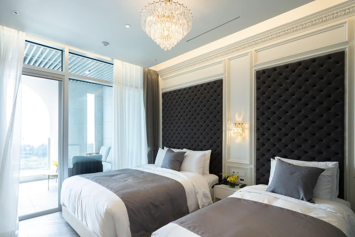 A modern hotel room with two beds, a large window, and a chandelier.