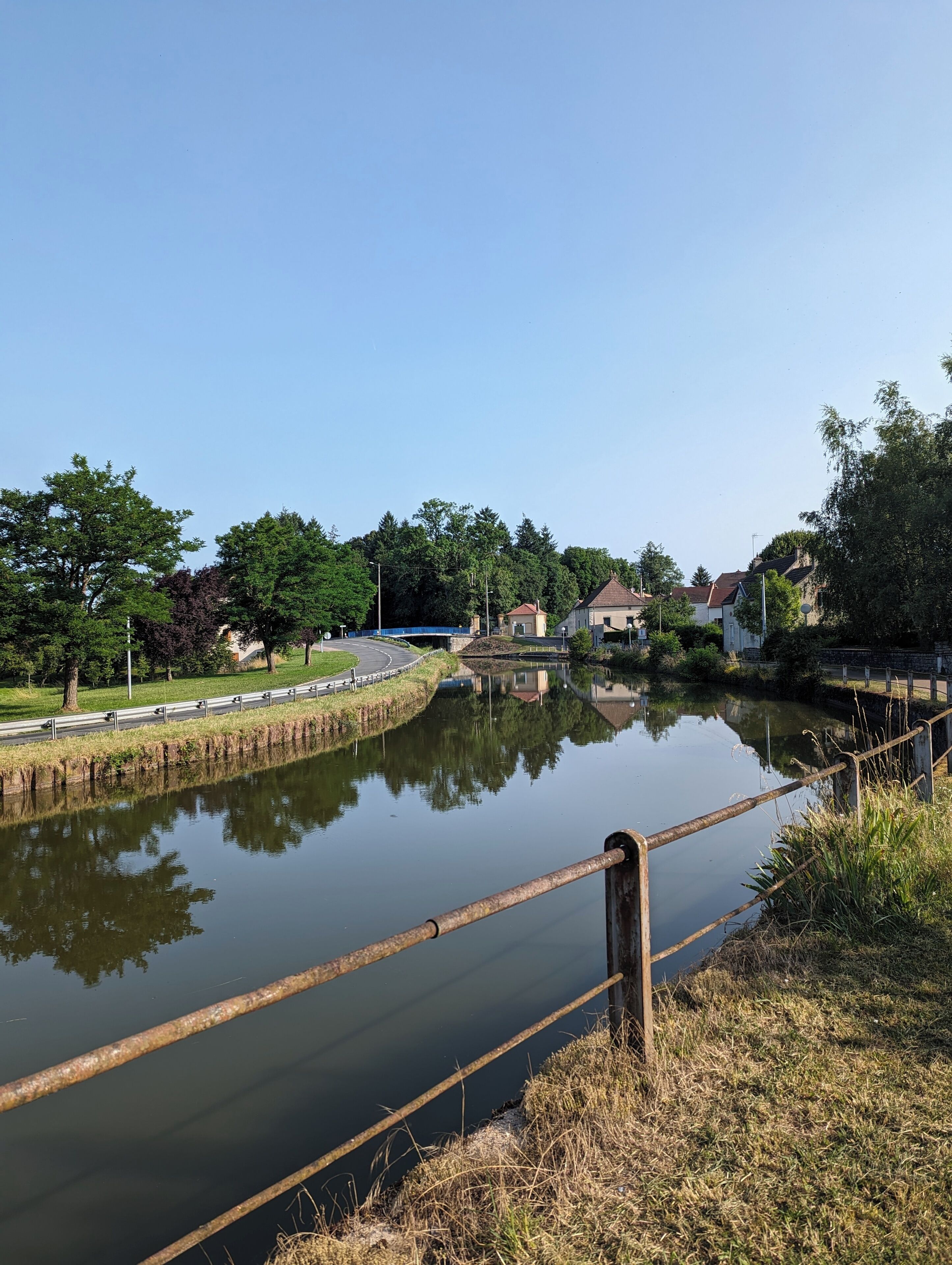 Een rustige rivier met een metalen hek, omgeven door groen en huizen.