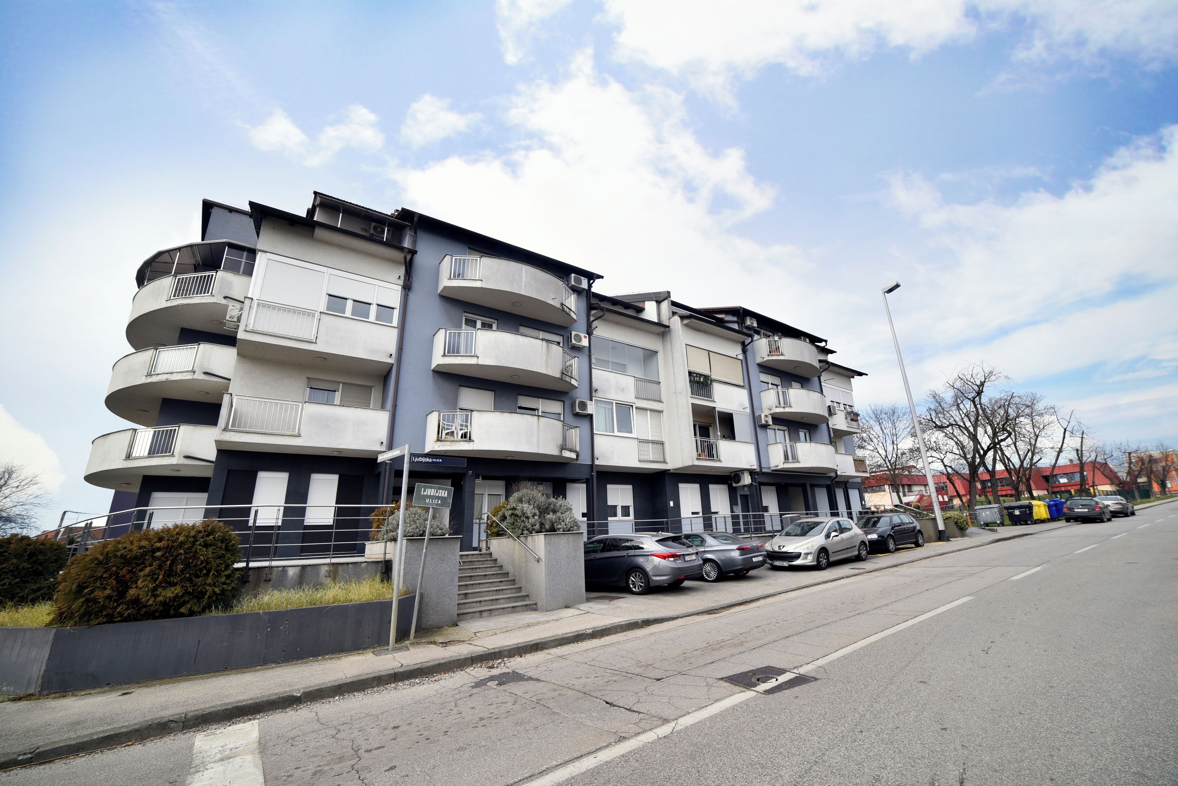 A multi-story residential building with balconies, a parking area, and a clear sky.