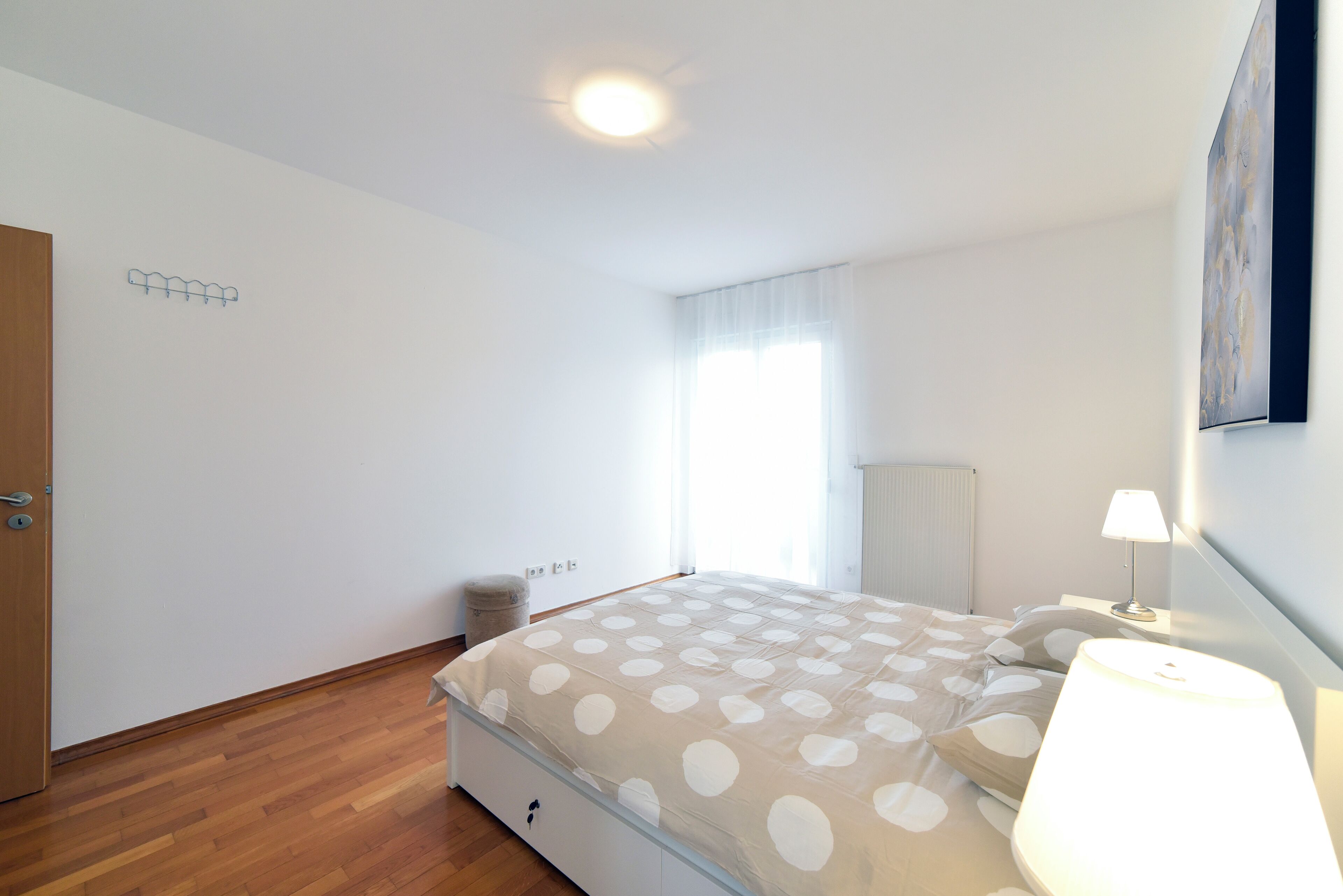A bedroom with a bed, a bedside lamp, a radiator, a window with curtains, and a door.