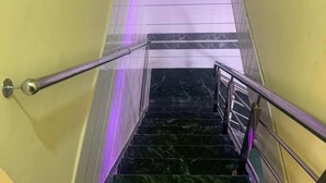 Una escalera con escalones de mármol y barandillas de metal, un extintor en la plataforma, y una tira de luz púrpura en la pared.