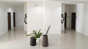 Un hall d’entrée spacieux et moderne, aux murs blancs et au sol carrelé, agrémenté de plantes en pot et de panneaux indicateurs pour les toilettes.