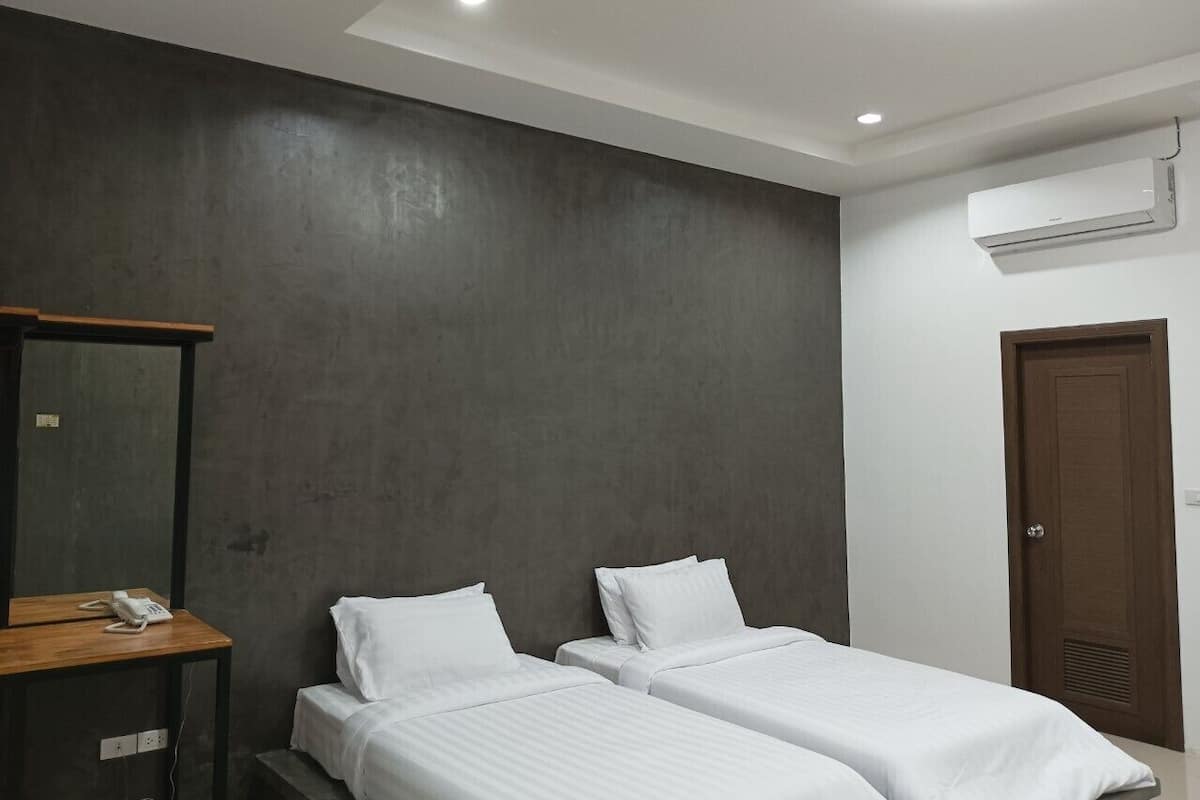Quarto de hotel com duas camas de solteiro, uma mesa de cabeceira de madeira com telefone, parede escura texturizada, teto branco e um aparelho de ar condicionado.