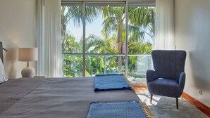 A bedroom with a large bed, a chair, and a view of palm trees through a window.