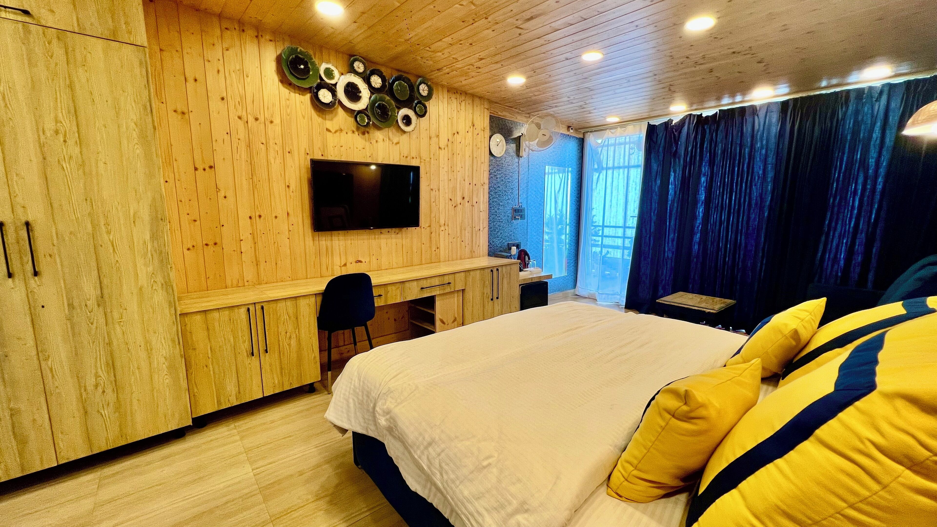A bedroom with a wooden wall, a bed with yellow pillows, a desk, and a TV.