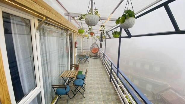 A balcony with hanging planters, a wooden table, blue chairs, and a hanging orange chair. - Clarence Cottage (Dehradun)