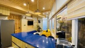 A modern kitchen with a blue countertop, wooden cabinets, and a built-in microwave. There is a yellow sofa and a window with a view of the outdoors. - Clarence Cottage (Dehradun)