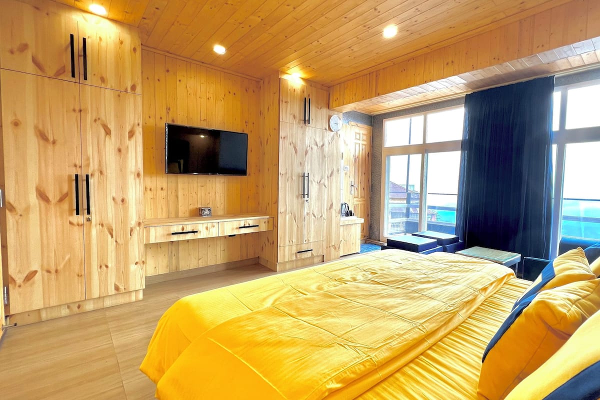 A wooden cabin bedroom with a bed, a TV, a window with curtains, and a view of the outside.
