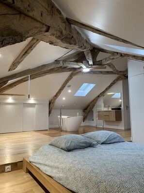 Une chambre avec un plafond en pente, des poutres apparentes, un puits de lumière, un grand lit et une salle de bain avec baignoire.