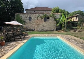 Une piscine rectangulaire bordée de pierres, entourée d’un muret en pierre et d’une pelouse. On y trouve des chaises longues et une table avec un parasol.