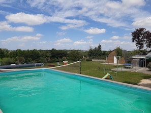 A swimming pool with a netted area, surrounded by a grassy yard, a small shed, and a wooden structure.