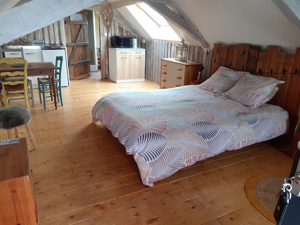 A cozy attic bedroom with a wooden floor, a bed with a patterned bedspread, a desk with a computer, and a kitchenette in the background.