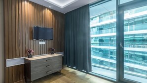 A modern hotel room with a wooden panel wall, a flat-screen TV, a wooden dresser, and a large sliding glass door leading to a balcony with a view of a building. - GLOBALSTAY Batumi View Apartments. Beach (Batumi)