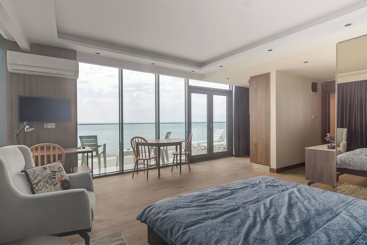 A modern hotel room with a large bed, a seating area with a table and chairs, a TV, and a balcony with a view of the sea.