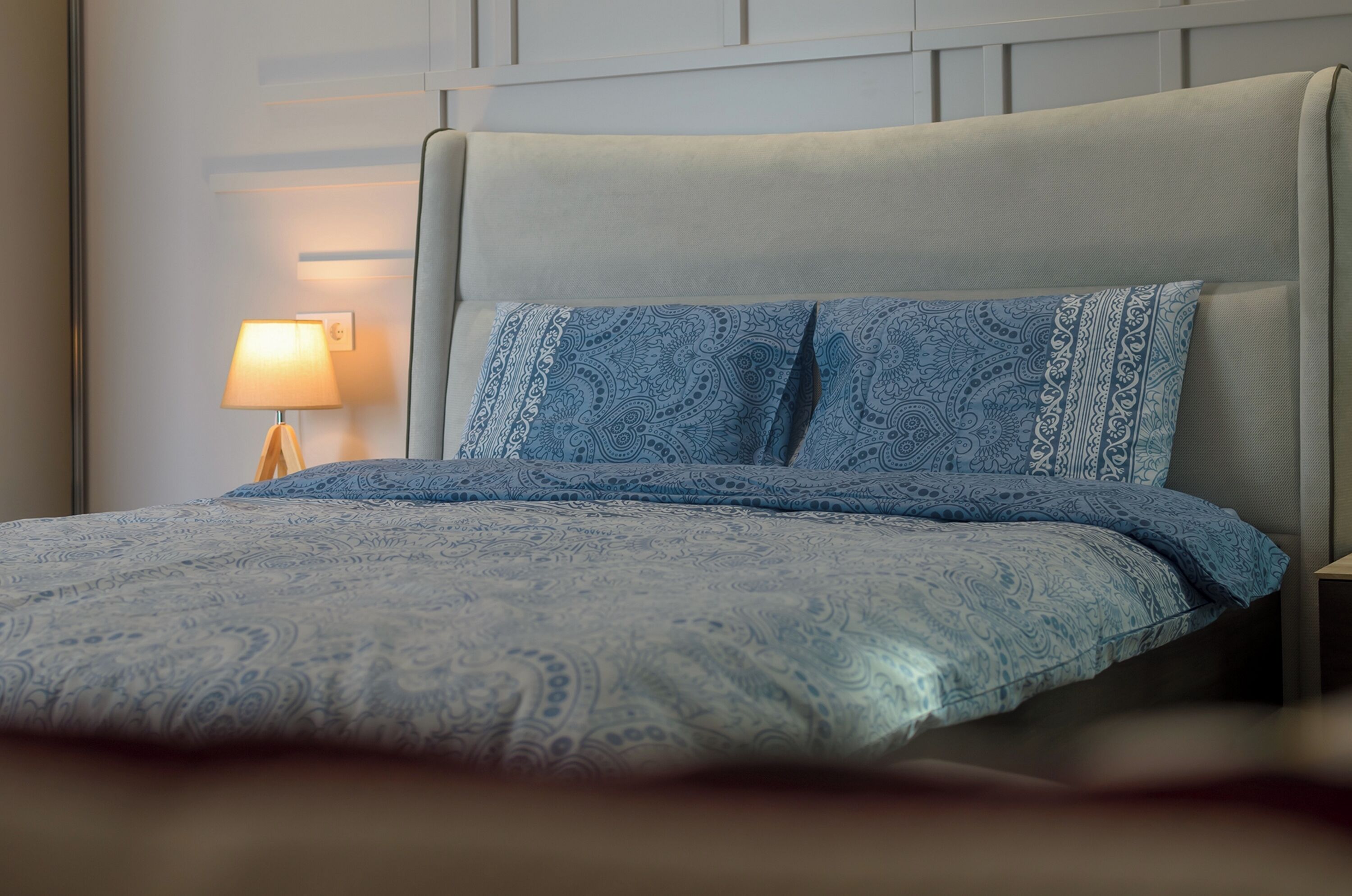 Una cama bien hecha con una colcha estampada y almohadas, una lámpara de noche y un panel en la pared.