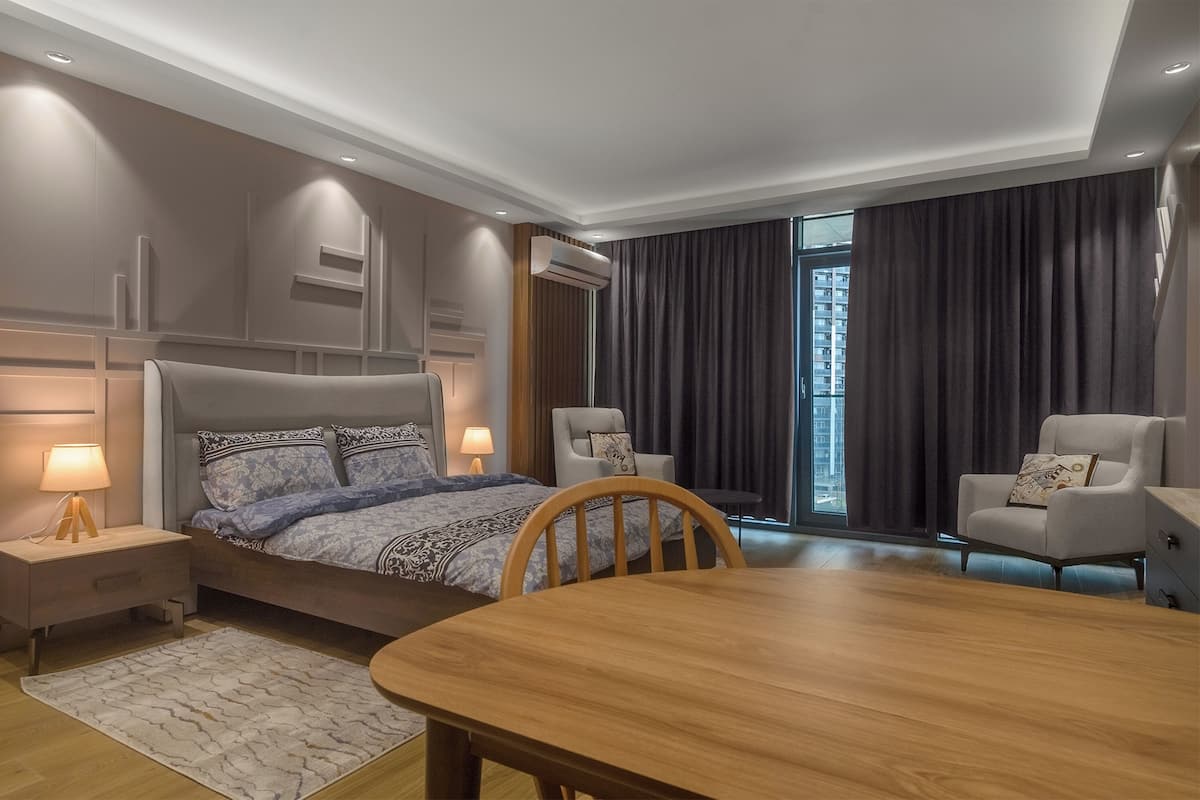 A modern hotel room with a large bed, two armchairs, a wooden dining table, and a large window with curtains.