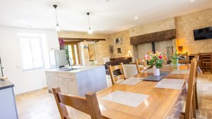 A spacious dining area with a long wooden table and chairs, a kitchen in the background, and a stone wall. - Charming Country House in Tourtoirac, Pets Allowed (Tourtoirac)