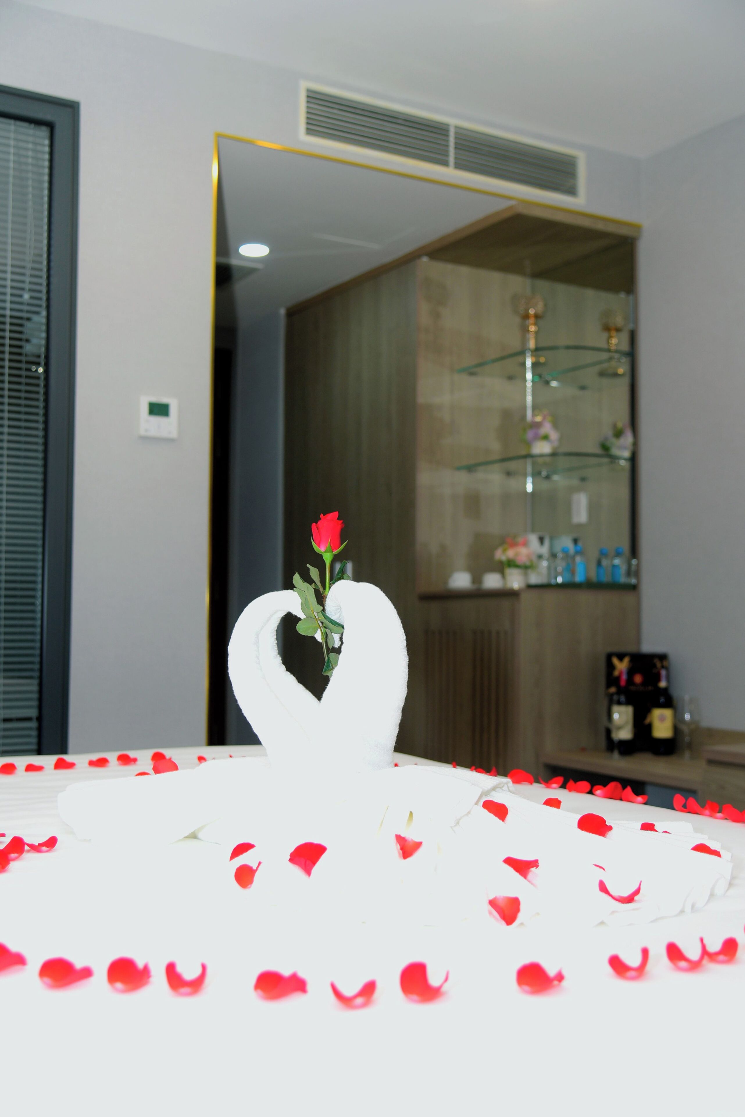 Una habitación con una cama cubierta de pétalos de rosa y una sola rosa roja en un florero con forma de corazón.