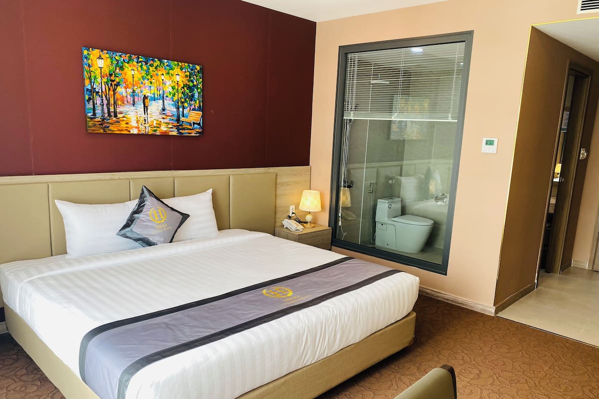 Una habitación de hotel con cama, mesita de noche, lámpara, un cuadro y la puerta del baño.