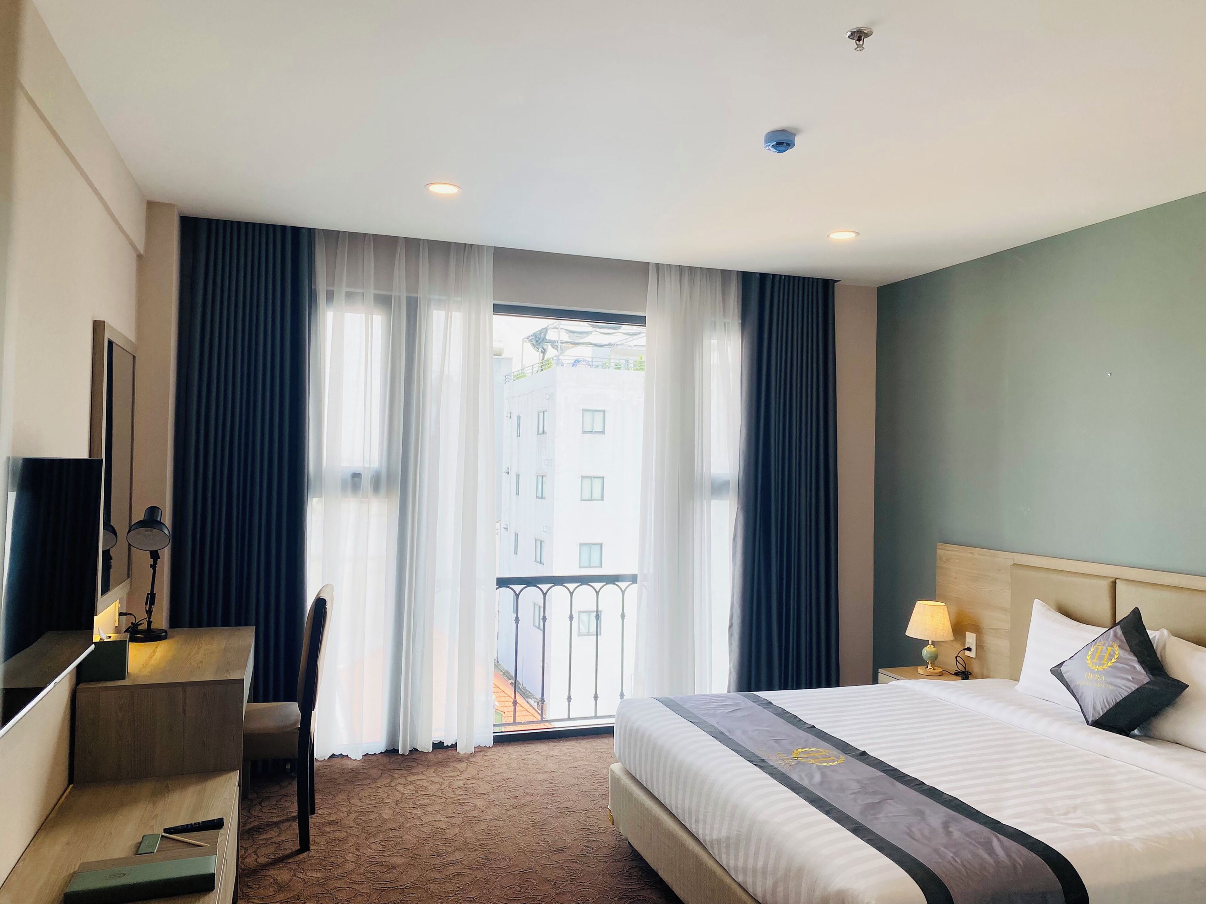 A hotel room with a bed, desk, chair, and a view of buildings through the window.