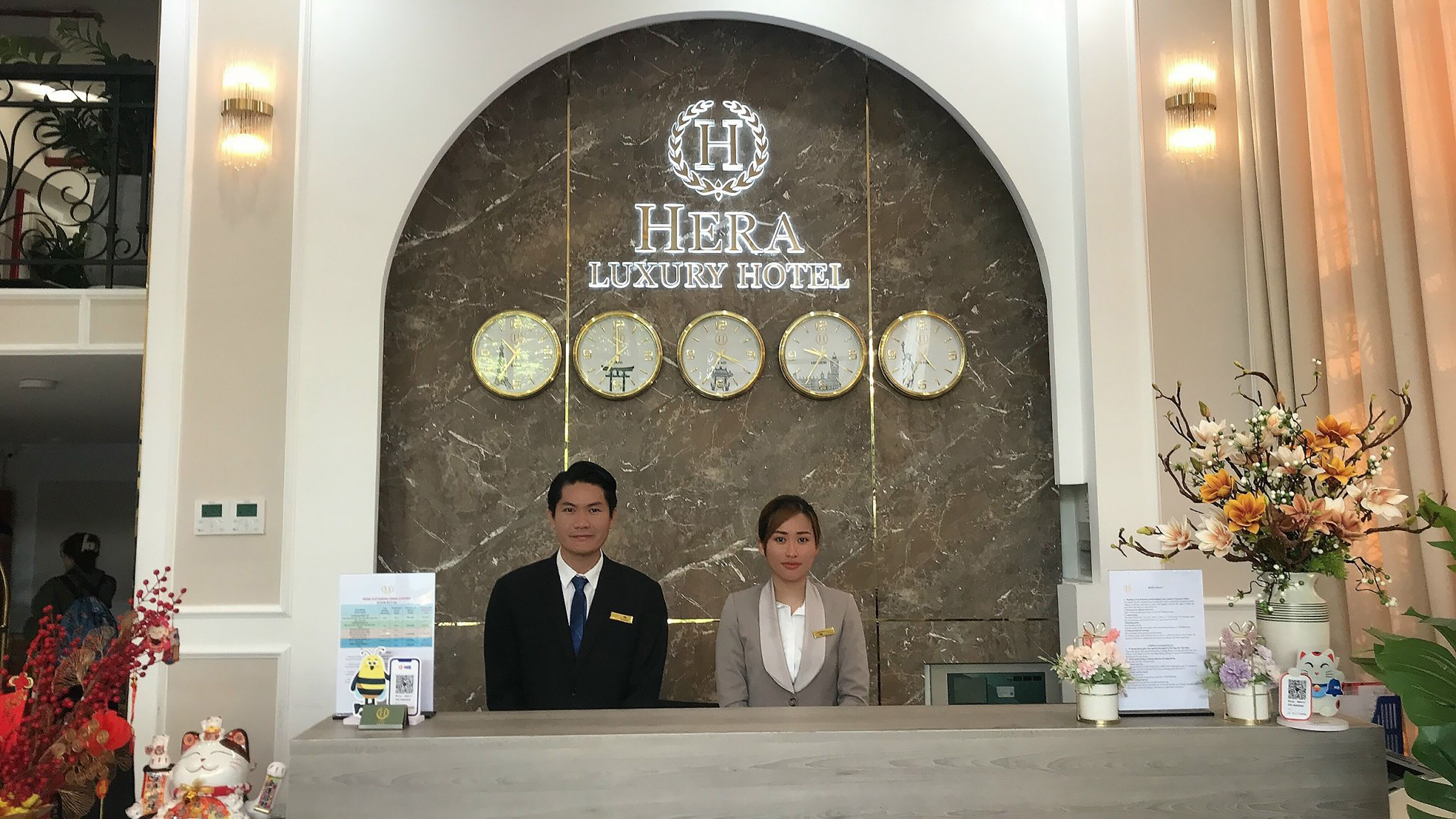 A reception desk at Hera Luxury Hotel with a marble wall, clocks, and a sign.