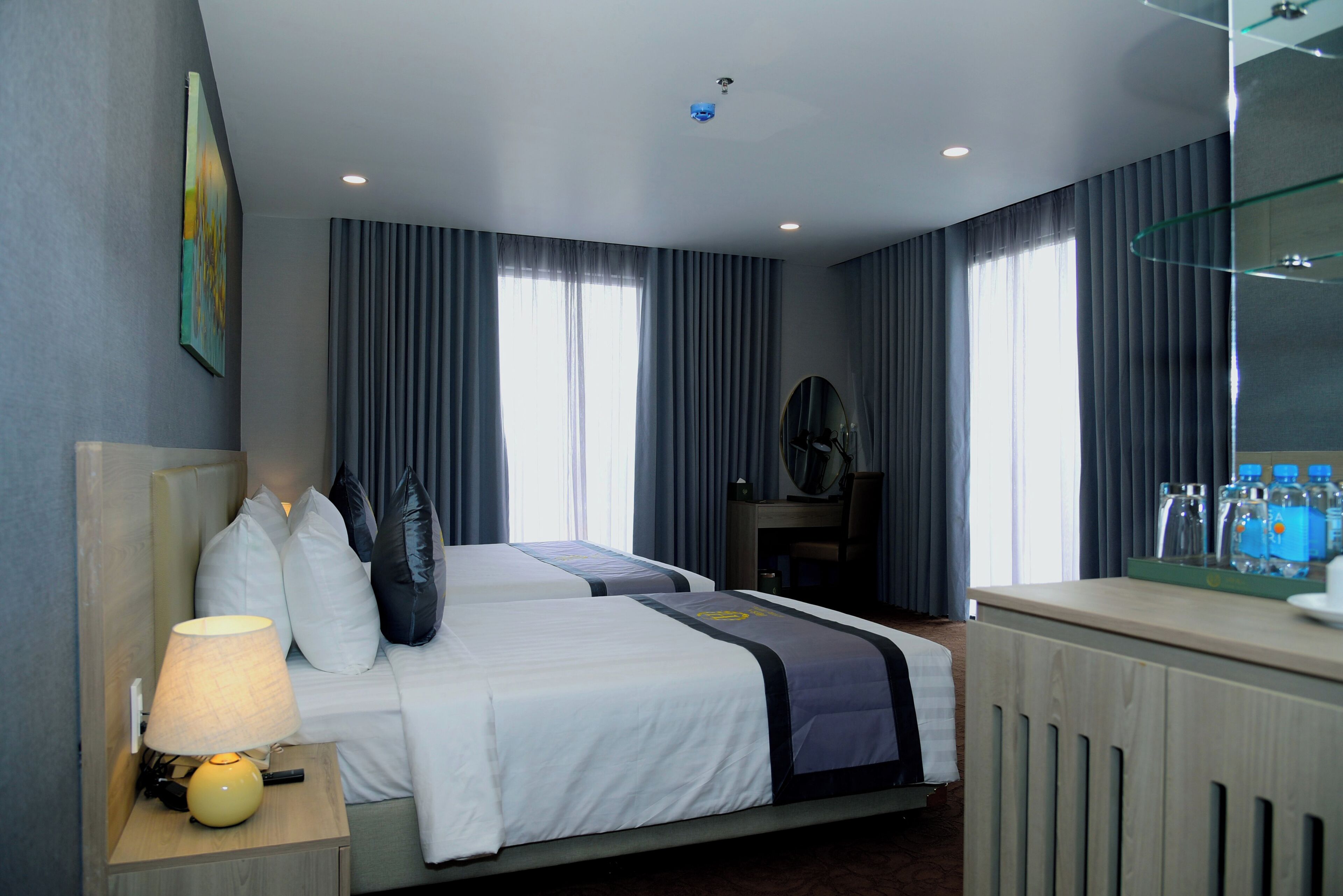 A hotel room with two beds, a bedside table with a lamp, a mirror, a dresser, and a window with curtains.
