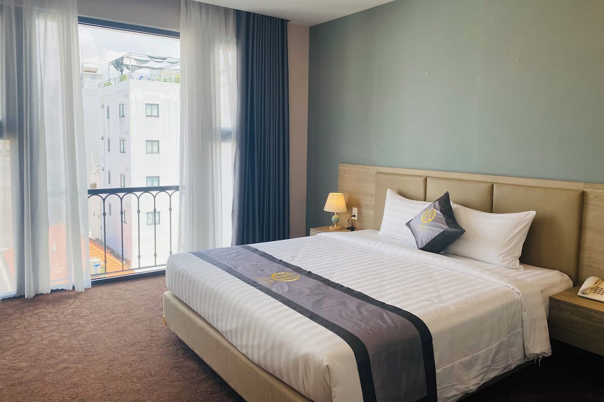 Habitación de hotel con una cama grande, una mesita de noche con lámpara y vistas a edificios a través del ventanal.