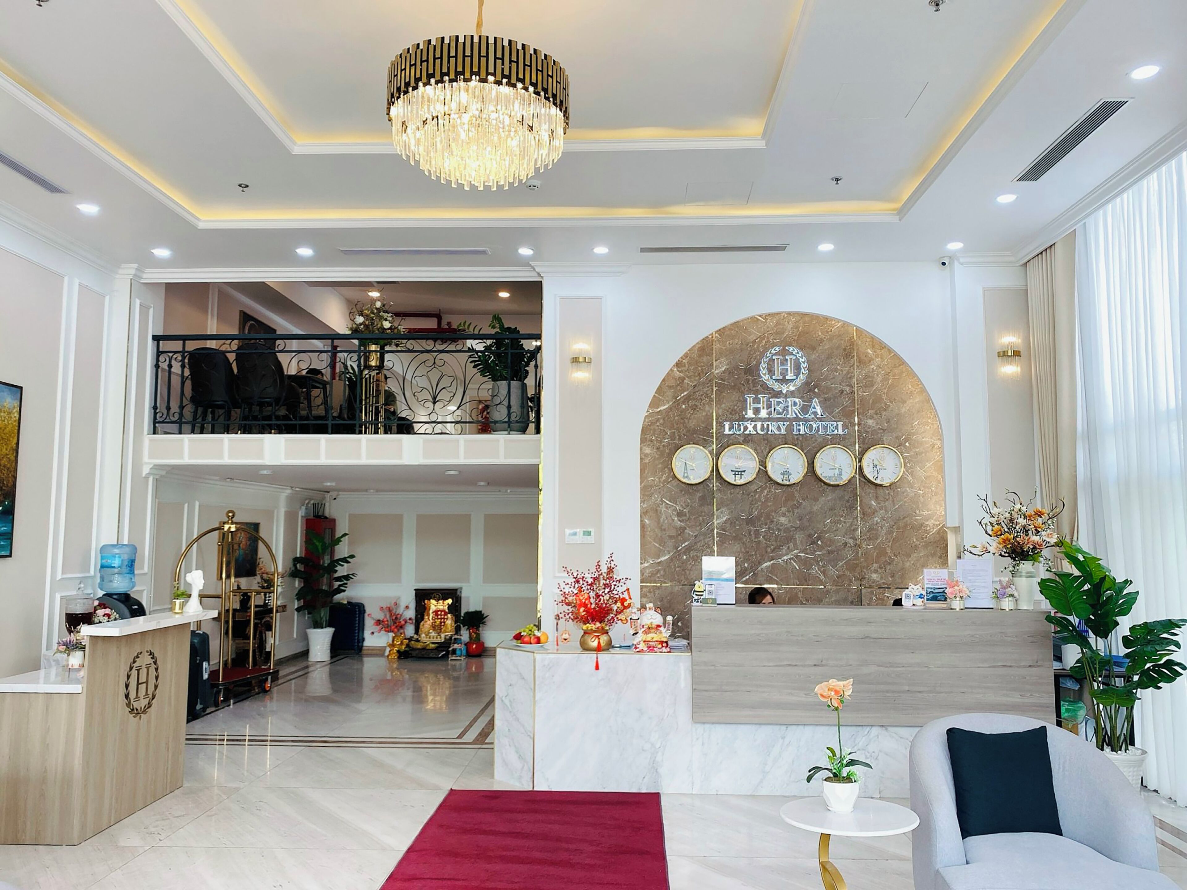 A hotel lobby with a reception desk, a chandelier, a marble wall with the hotel name, and a balcony with chairs.