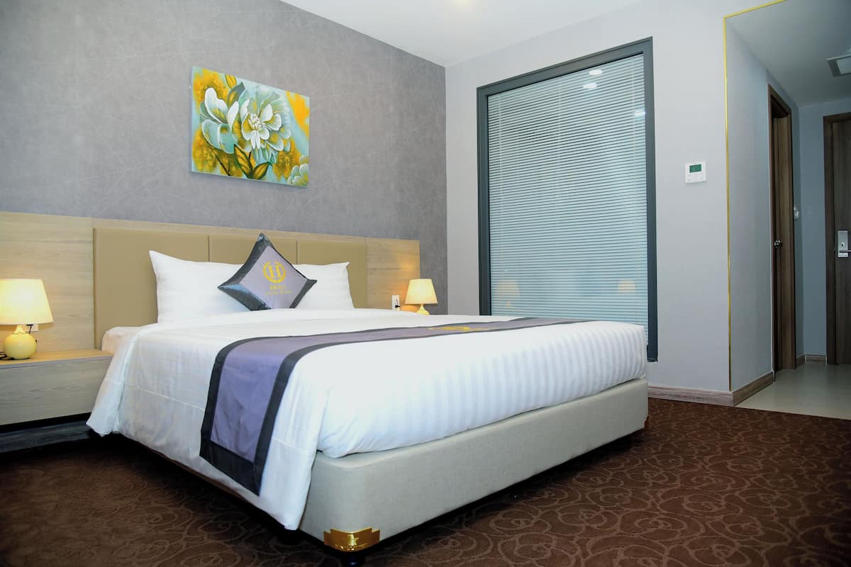 Habitación de hotel con una cama grande, una mesita de noche con lámpara, un cuadro en la pared y una ventana con persianas.