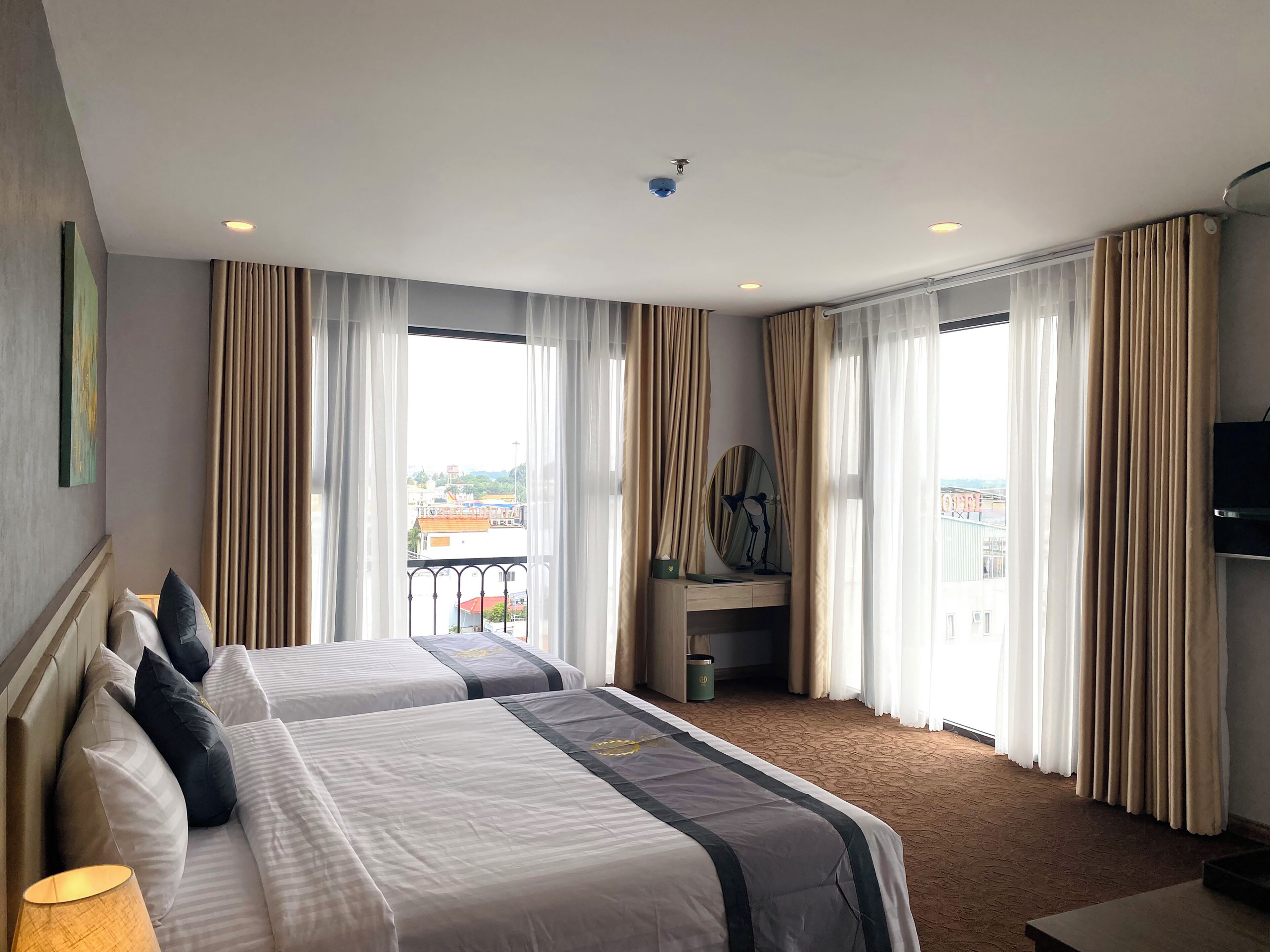 A hotel room with two beds, a desk, a chair, a TV, and large windows with curtains.
