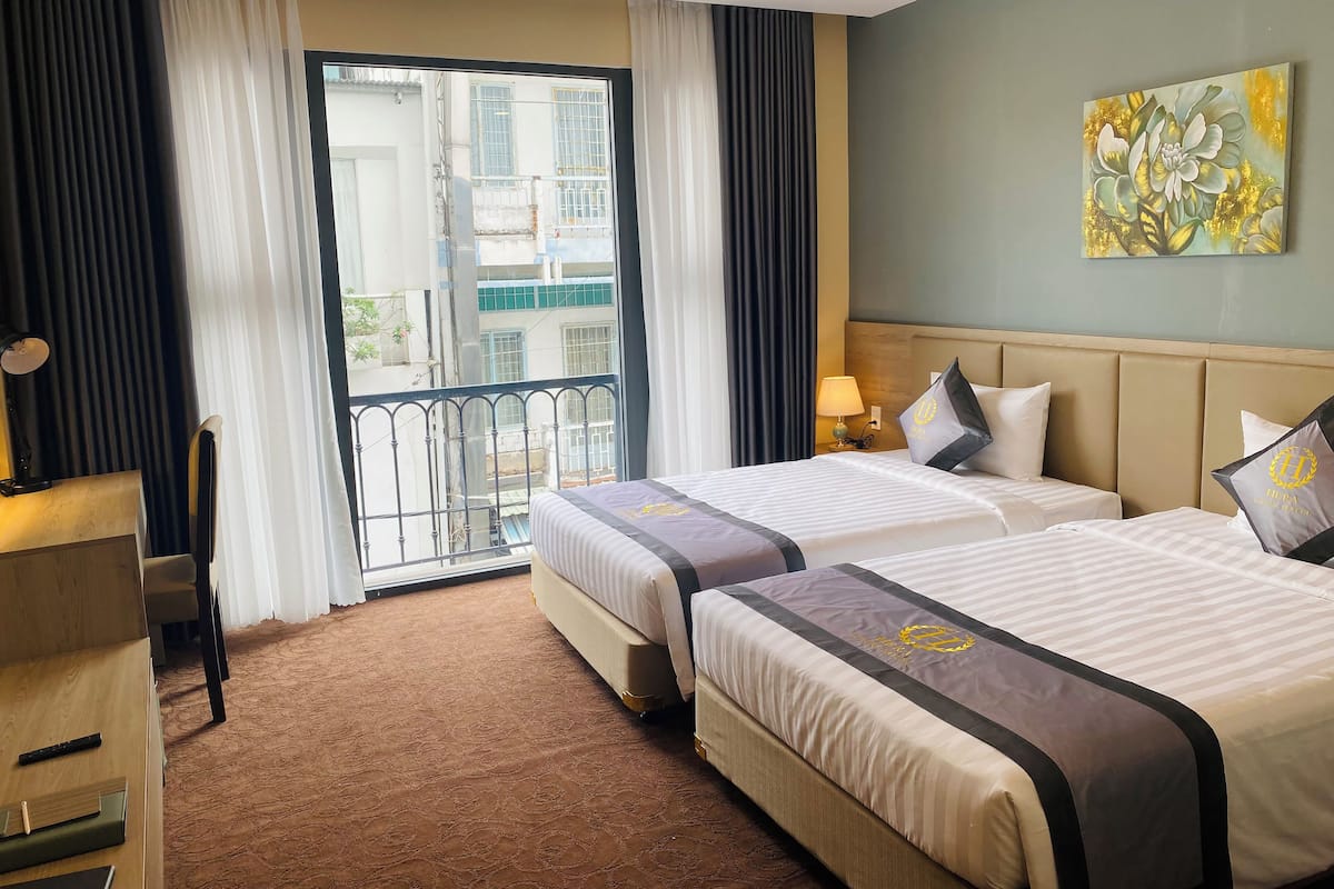 Habitación de hotel con dos camas, un escritorio, una silla, un balcón con vistas a edificios y un cuadro en la pared.