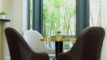 A modern dining area with two brown chairs and a small table, large windows with a view of bamboo outside, and a ceiling-mounted air conditioner.