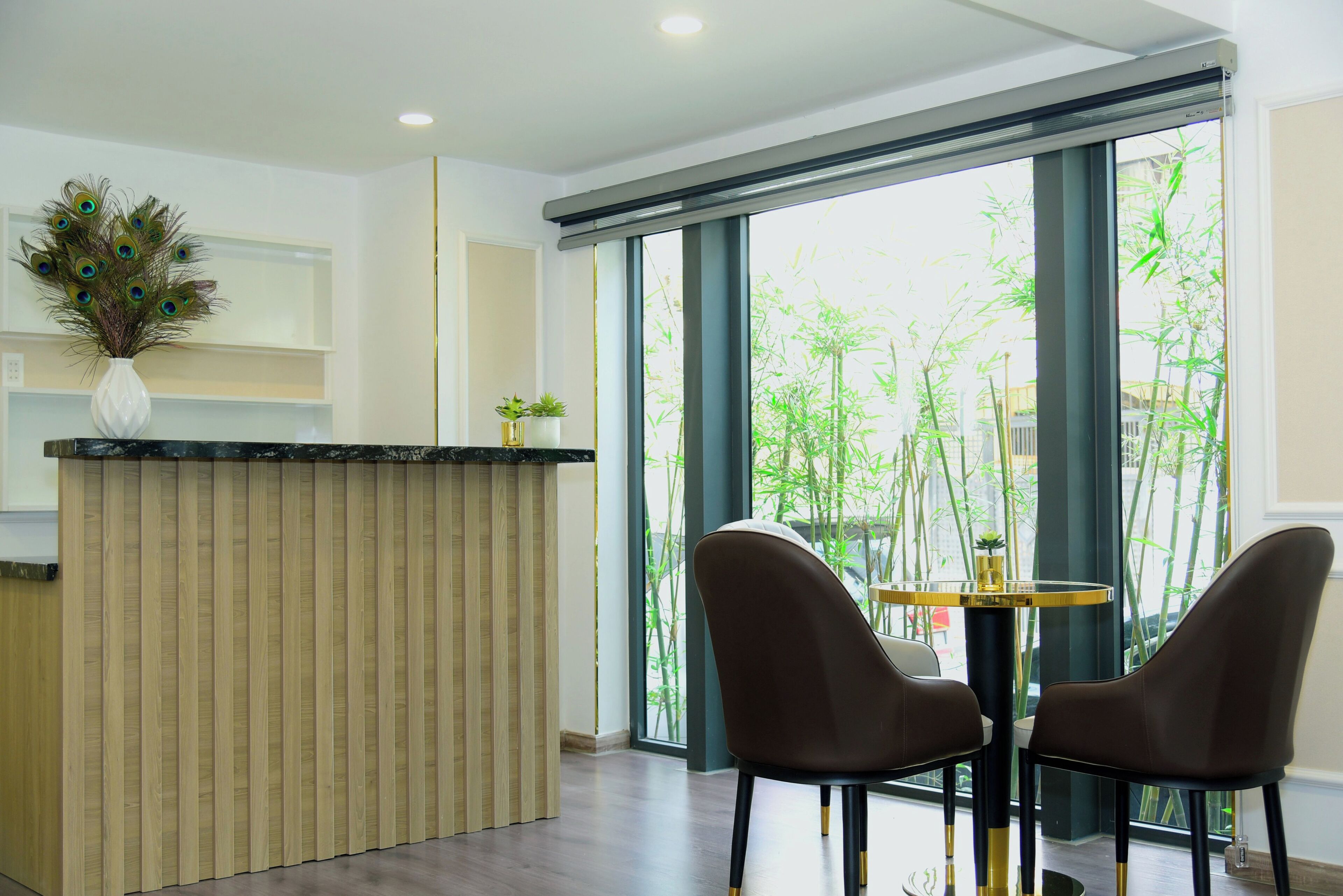 A reception area with a wooden reception desk, two brown chairs, a small table with a plant, and large windows with a view of bamboo outside.