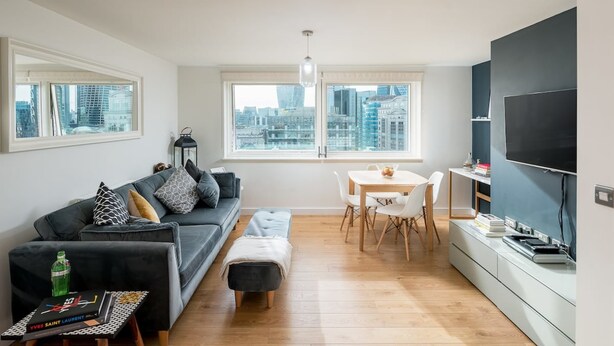 A modern living room with a sofa, ottoman, dining table, and chairs. There is a large window with a city view, a flat-screen TV, and a bookshelf.