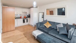 A modern living room with a grey sofa, a wooden floor, and a kitchen in the background.