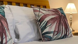 Un lit avec une tête de lit en bois et deux oreillers, un blanc et l’autre orné d’un motif de feuilles tropicales. Une lampe avec un abat-jour blanc est visible sur le côté.