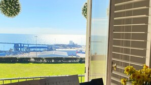 A room with a large window overlooking a port with cranes and ships, a green lawn, and a chair with yellow flowers. - Recently refurbished and sleeping up to 4 guests, this stylish apartment offers stunning sea views, modern interiors, a fully equipped kitchen and a prime central location. Just moments from Ramsgate's Royal Harbour, beach, cafés and restaurants. (Kent)