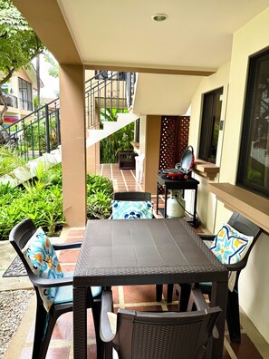An outdoor patio area with a dining table and chairs, surrounded by greenery and a view of a building with a balcony.
